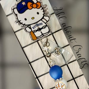 May include: A badge reel featuring a Hello Kitty character wearing a blue and orange Houston Astros baseball cap and uniform, holding a bat. The character has a glittery border and is attached to a retractable cord with blue and white beads.
