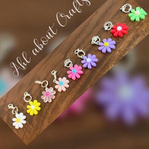 May include: A collection of colorful daisy flower charms with silver-tone clasps. The flowers are in various colors, including white, yellow, pink, purple, and green. The charms are arranged on a wooden surface, with the text "Ahl about Crafts" in the upper left corner.