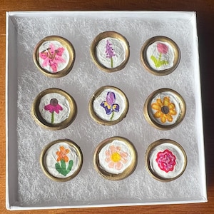May include: A white box with a white background containing nine round, gold-rimmed, white paper circles with pressed flowers inside. The flowers are a variety of colors, including pink, purple, yellow, and orange.