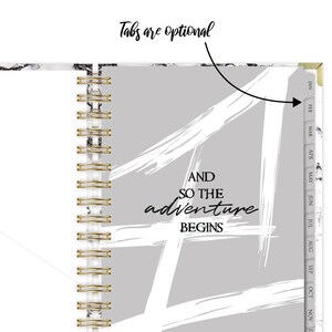 Może przedstawiać: Szary i biały planer z marmurową okładką i złotym spiralnym oprawieniem. Okładka ma biały wz&oacute;r z pociągnięć pędzlem i tekst "AND SO THE adventure BEGINS". Planer ma zakładki na każdy miesiąc roku.