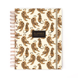 May include: A cream-colored planner with a gold spiral binding and a gold ribbon bookmark. The cover features a repeating pattern of brown tigers on a cream background. The planner is labeled "PLANNER 2023-2024".
