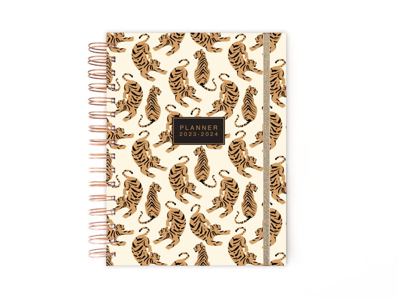 May include: A cream-colored planner with a gold spiral binding and a gold ribbon bookmark. The cover features a repeating pattern of brown tigers on a cream background. The planner is labeled "PLANNER 2023-2024".