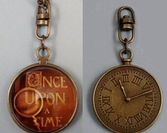 May include: A bronze-colored pendant necklace with a round charm. One side features the words "ONCE UPON A TIME" in a vintage font with a magnifying glass illustration. The other side is a clock face with Roman numerals. The necklace has a chain and a clasp.
