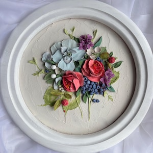 May include: A circular, white-framed relief sculpture of a floral arrangement. The artwork depicts red roses, blue hydrangeas, purple flowers, and blueberries. Green leaves and white berries are set against a textured, off-white background, within a white frame.