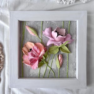 May include: A framed floral artwork featuring two open flowers and two buds in shades of pink and peach, with green leaves and stems. The flowers are set against a textured gray background with vertical green lines, all within a white wooden frame.