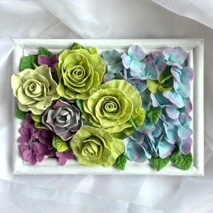 May include: A rectangular white framed floral arrangement featuring green roses, purple hydrangeas, and blue hydrangeas. The flowers are arranged in a dense, textured composition, with green leaves interspersed throughout.
