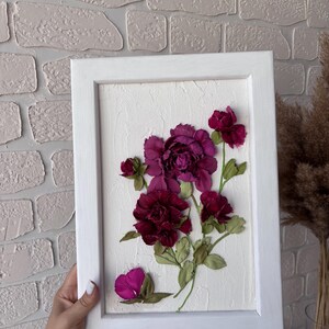 May include: A framed floral artwork with deep burgundy and magenta flowers and green leaves. The flowers are set against a textured white background within a white wooden frame. The artwork is held by a person.
