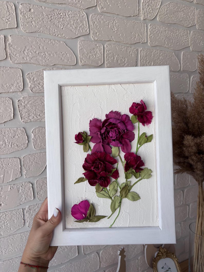 May include: A framed floral artwork with deep burgundy and magenta flowers and green leaves. The flowers are set against a textured white background within a white wooden frame. The artwork is held by a person.