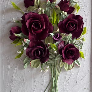 May include: A bouquet of burgundy roses with green leaves and stems, set against a textured white background. The handmade floral arrangement features detailed petals and small white accents, creating a three-dimensional effect.