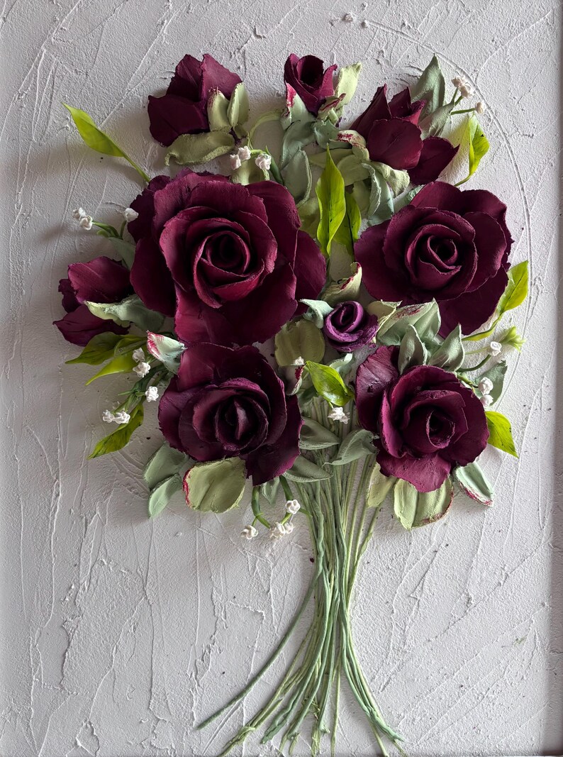 May include: A bouquet of burgundy roses with green leaves and stems, set against a textured white background. The handmade floral arrangement features detailed petals and small white accents, creating a three-dimensional effect.