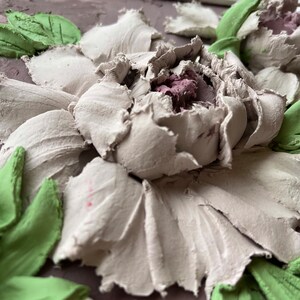 May include: Close-up of a 3D floral wall art piece with a white flower with pink center and green leaves. The flower is textured and appears to be made of plaster or clay.