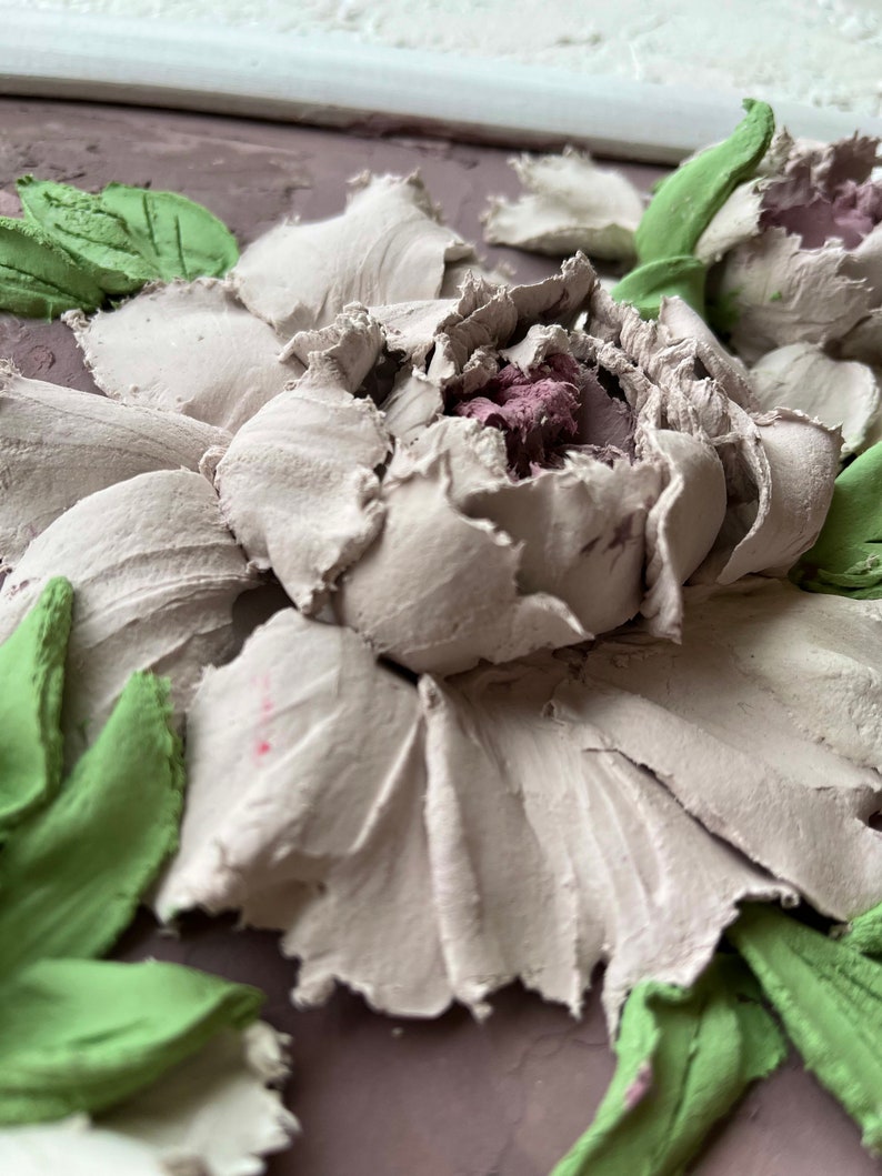 May include: Close-up of a 3D floral wall art piece with a white flower with pink center and green leaves. The flower is textured and appears to be made of plaster or clay.