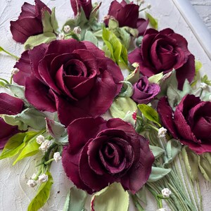 May include: A framed floral artwork featuring a bouquet of burgundy roses, buds, and green leaves. The roses have textured petals and are surrounded by small white flowers. The artwork is set against a textured white background.