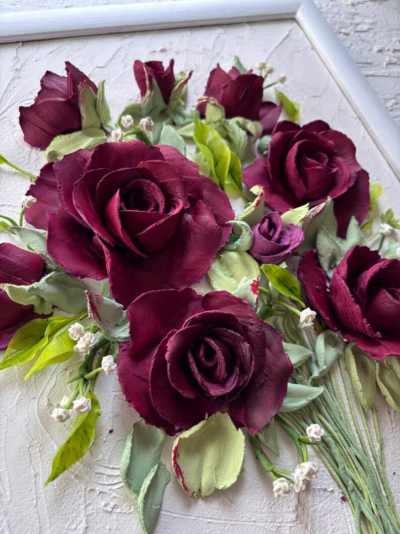 May include: A framed floral artwork featuring a bouquet of burgundy roses, buds, and green leaves. The roses have textured petals and are surrounded by small white flowers. The artwork is set against a textured white background.