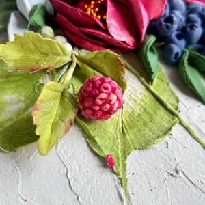 May include: A close-up of a detailed floral arrangement featuring a vibrant red raspberry, green leaves, and a deep red flower. The arrangement includes blueberries and white accents, all set against a textured white background.