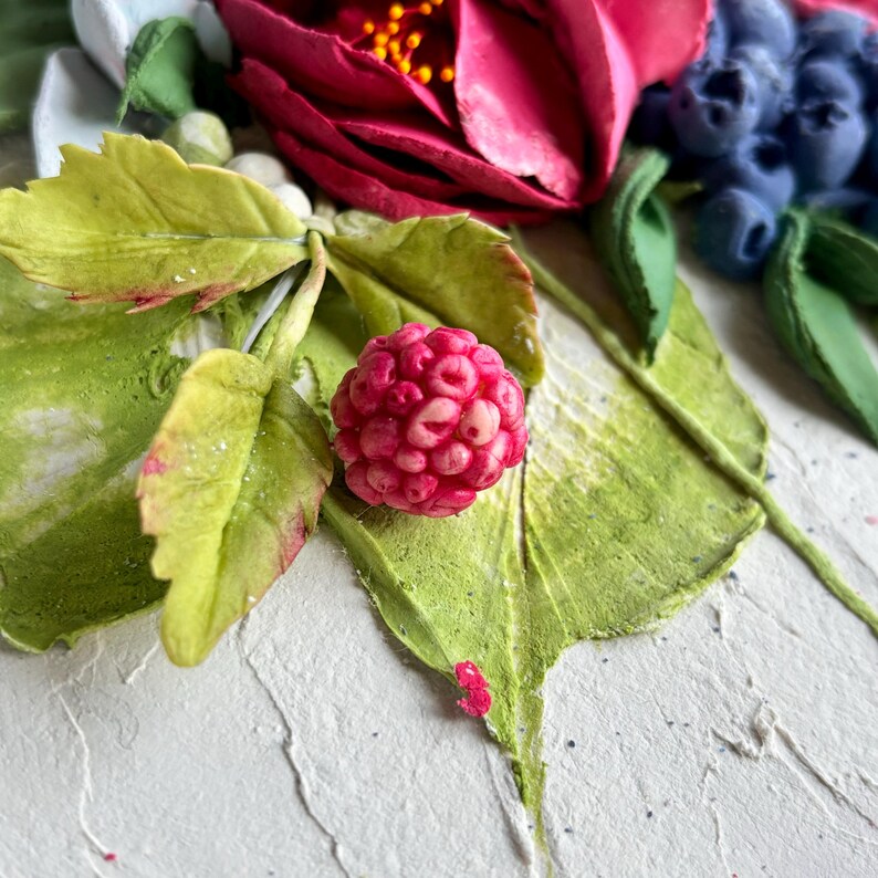 May include: A close-up of a detailed floral arrangement featuring a vibrant red raspberry, green leaves, and a deep red flower. The arrangement includes blueberries and white accents, all set against a textured white background.