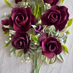 May include: A bouquet of burgundy roses with green leaves and small white flowers. The roses are the focal point, with detailed petals and a textured appearance. The bouquet is tied with green stems, set against a textured white background.