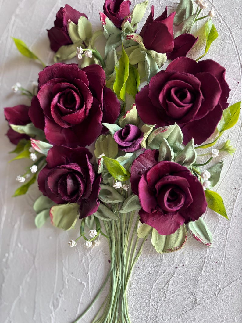 May include: A bouquet of burgundy roses with green leaves and small white flowers. The roses are the focal point, with detailed petals and a textured appearance. The bouquet is tied with green stems, set against a textured white background.