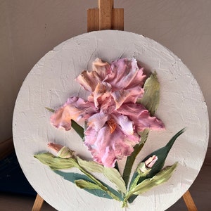 K&ouml;nnte beinhalten: Ein rundes Leinwandbild mit einer rosa und wei&szlig;en Irisbl&uuml;te mit gr&uuml;nen Bl&auml;ttern. Das Bild steht auf einem Staffelei.