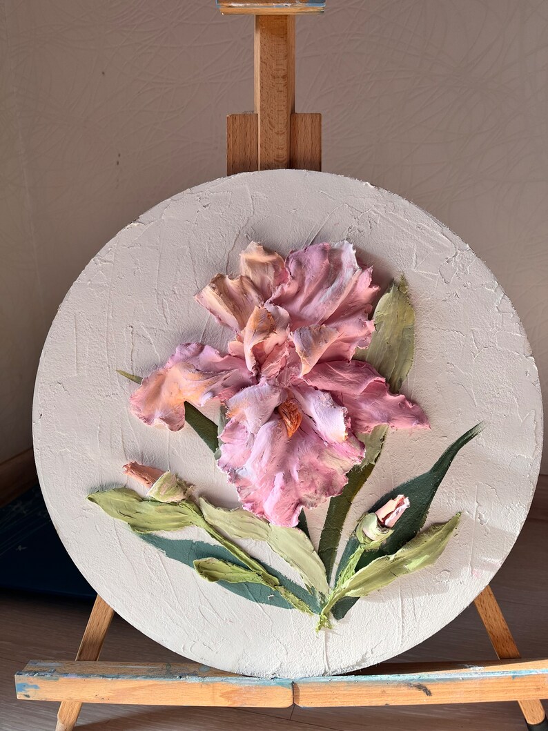 K&ouml;nnte beinhalten: Ein rundes Leinwandbild mit einer rosa und wei&szlig;en Irisbl&uuml;te mit gr&uuml;nen Bl&auml;ttern. Das Bild steht auf einem Staffelei.