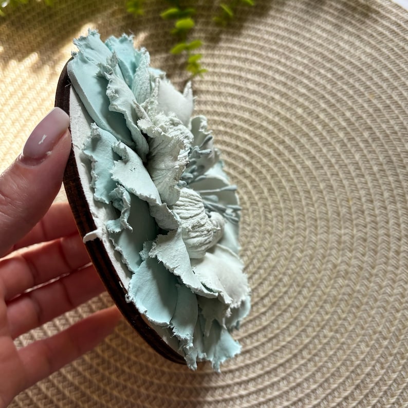 以下が含まれることがあります： 薄い青色の布でできた花が、丸い木製の土台に付けられています。花は、層状でフリルのある外観をしています。