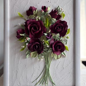 May include: A framed artwork featuring a bouquet of burgundy roses with green leaves and small white accents. The flowers are arranged in a textured, off-white background within a white frame. The roses are the focal point, with detailed petals.