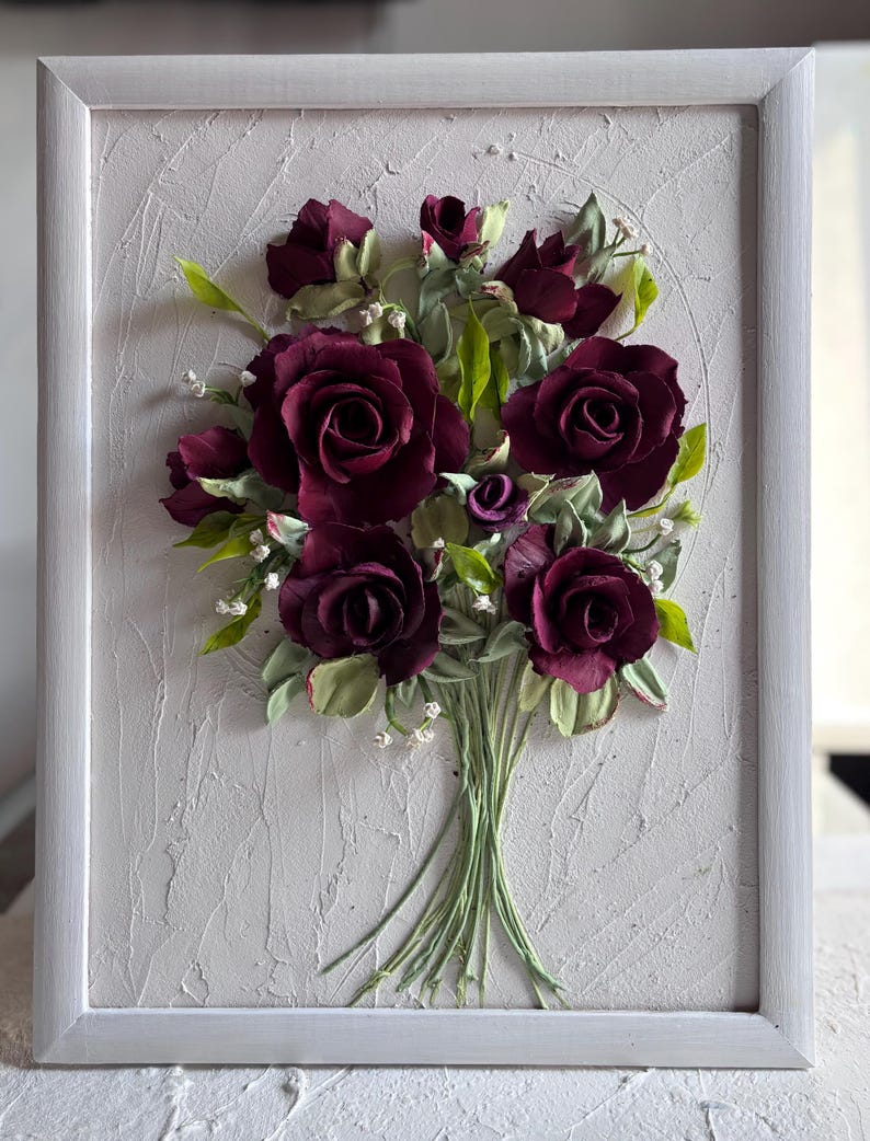 May include: A framed artwork featuring a bouquet of burgundy roses with green leaves and small white accents. The flowers are arranged in a textured, off-white background within a white frame. The roses are the focal point, with detailed petals.