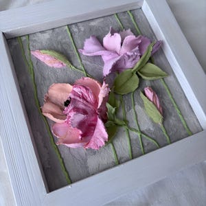 May include: A framed floral artwork featuring two large, textured flowers in shades of pink and purple, with green leaves and stems. The flowers are set against a gray background with vertical green lines, all within a white wooden frame.