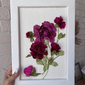 May include: A framed floral artwork featuring deep burgundy and magenta flowers with green leaves. The flowers are arranged on a textured white background within a white wooden frame. The artwork is held by a person.