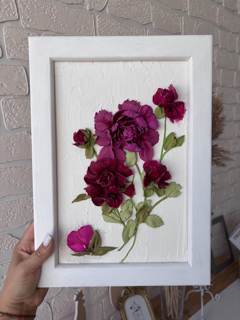 May include: A framed floral artwork featuring deep burgundy and magenta flowers with green leaves. The flowers are arranged on a textured white background within a white wooden frame. The artwork is held by a person.