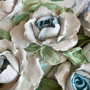 Puede incluir: Primer plano de una pintura texturizada de rosas blancas y azules con hojas verdes. Las rosas están pintadas en un estilo realista, con pinceladas visibles.