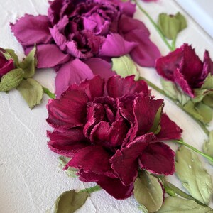 May include: A close-up of a floral artwork featuring textured, dimensional flowers in shades of deep red and purple. Green leaves and stems accent the blooms, set against a textured white background. The artwork evokes a sense of depth and natural beauty.