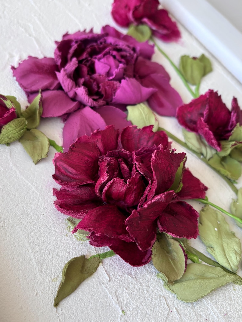 May include: A close-up of a floral artwork featuring textured, dimensional flowers in shades of deep red and purple. Green leaves and stems accent the blooms, set against a textured white background. The artwork evokes a sense of depth and natural beauty.