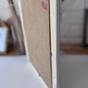May include: A light-coloured wooden picture frame. The reverse side is visible, showing a brown backing and a metal hanger. The frame has a simple design and a light wood finish.