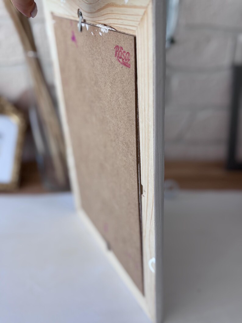 May include: A light-coloured wooden picture frame. The reverse side is visible, showing a brown backing and a metal hanger. The frame has a simple design and a light wood finish.