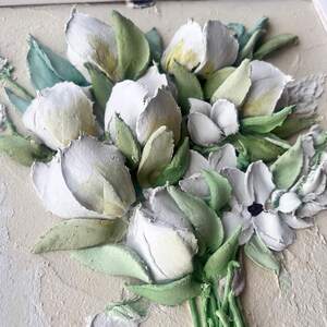 May include: A close-up of a white and green 3D floral bouquet. The flowers are made of textured paper and are arranged in a vase. The bouquet is set against a light beige background.