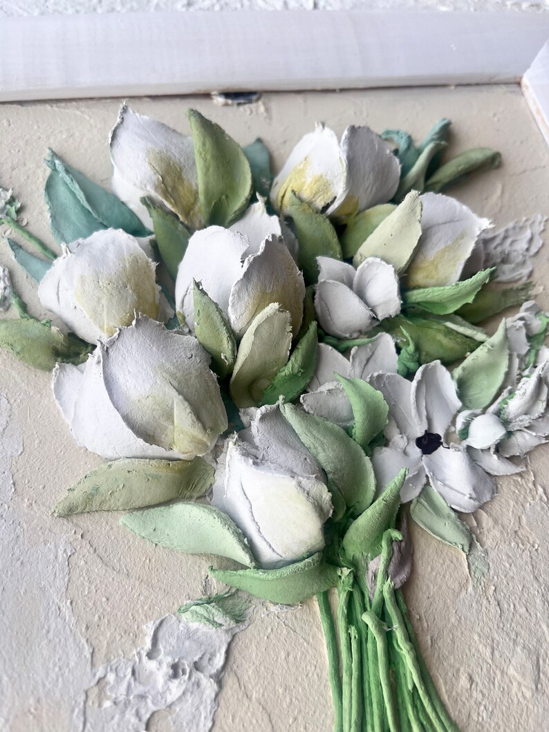 May include: A close-up of a white and green 3D floral bouquet. The flowers are made of textured paper and are arranged in a vase. The bouquet is set against a light beige background.