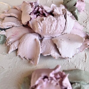 May include: Close-up of a textured, abstract floral artwork. The piece features a central flower with soft pink petals and a textured surface. Green and pink accents add depth and dimension to the piece. The artwork is on a light beige background.
