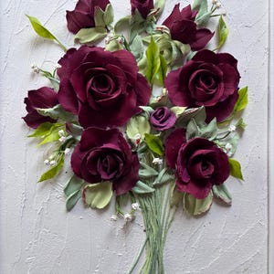 May include: A textured white framed artwork featuring a bouquet of burgundy roses with green leaves and stems. Small white flowers accent the arrangement. The roses are the focal point, with detailed petals.