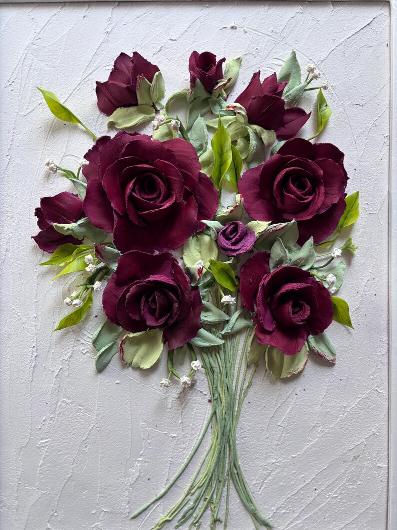 May include: A textured white framed artwork featuring a bouquet of burgundy roses with green leaves and stems. Small white flowers accent the arrangement. The roses are the focal point, with detailed petals.