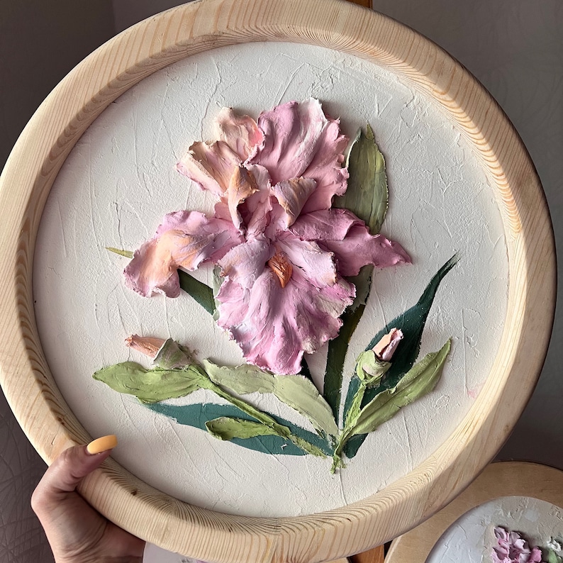 K&ouml;nnte beinhalten: Eine rosa und wei&szlig;e Iris-Blume mit gr&uuml;nen Bl&auml;ttern ist in 3D-Relief auf einer runden Holzplatte dargestellt.