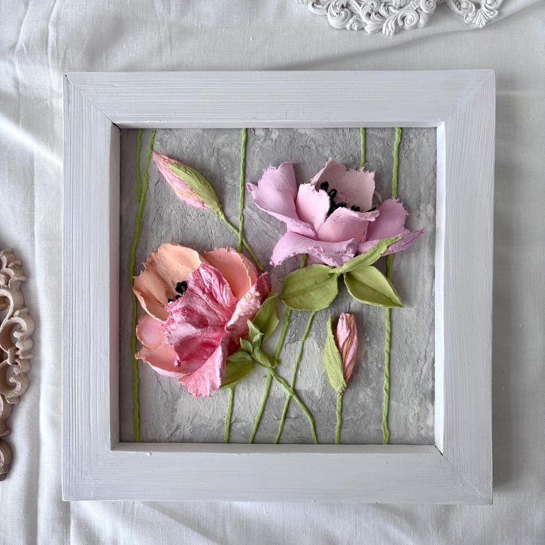 May include: A framed floral artwork featuring two open flowers and two buds in shades of pink and peach, with green leaves and stems. The flowers are set against a textured gray background with vertical green lines, all within a white wooden frame.