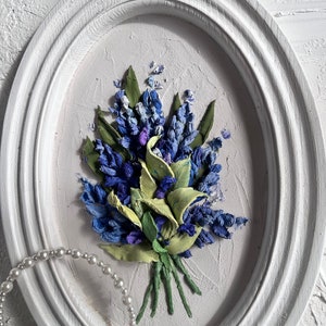May include: A white oval frame with a 3D floral painting of blue and green flowers. The painting is on a white background.