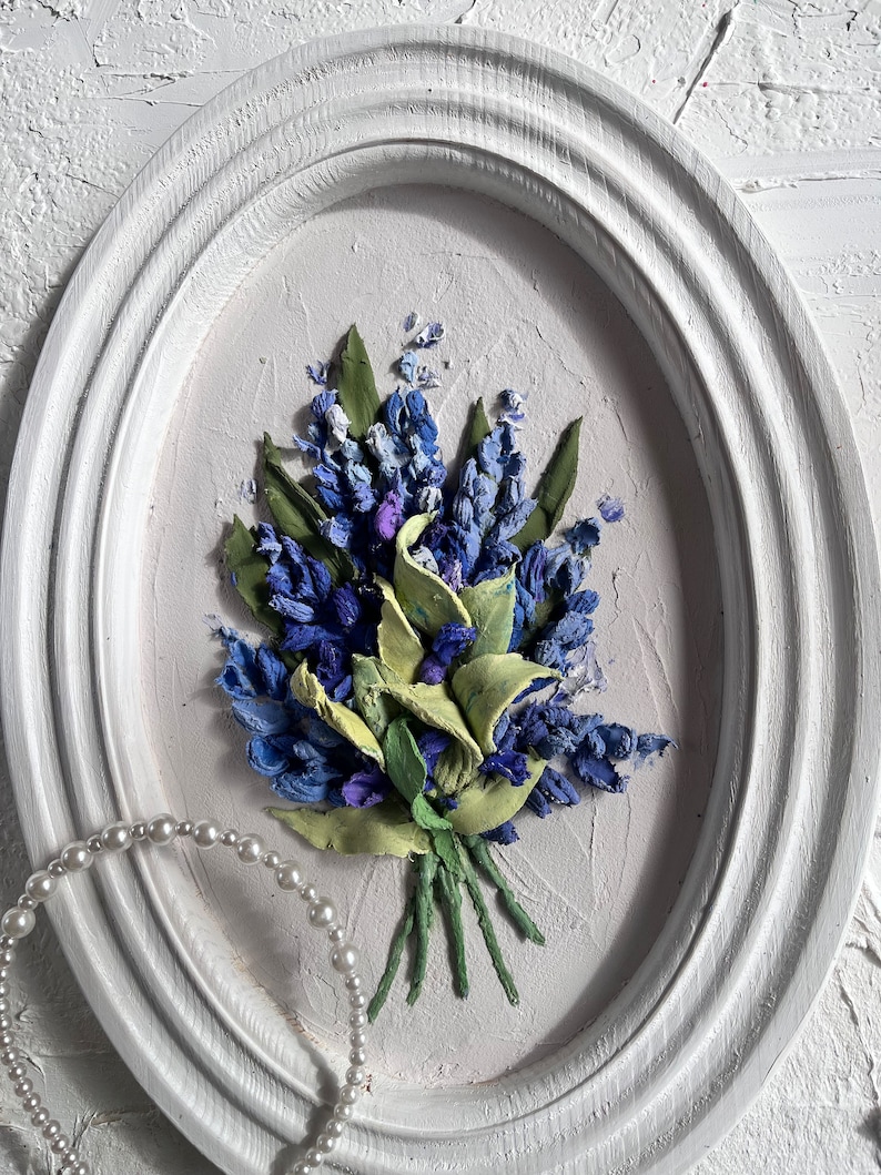May include: A white oval frame with a 3D floral painting of blue and green flowers. The painting is on a white background.