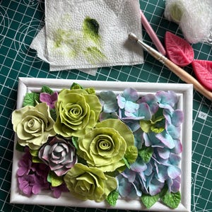 May include: A white framed floral arrangement featuring sculpted roses and hydrangeas in shades of green, purple, and blue. The flowers are arranged in a rectangular shape, creating a decorative piece. The background includes art supplies.