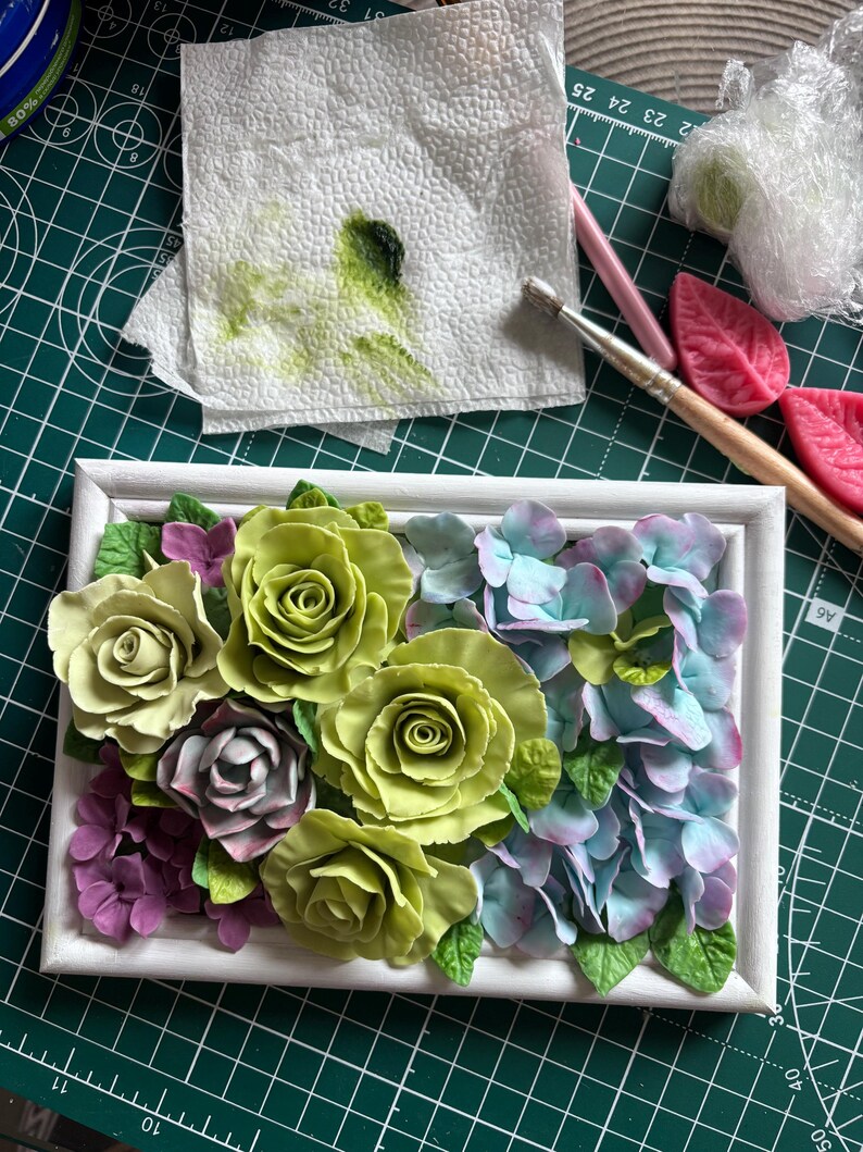 May include: A white framed floral arrangement featuring sculpted roses and hydrangeas in shades of green, purple, and blue. The flowers are arranged in a rectangular shape, creating a decorative piece. The background includes art supplies.