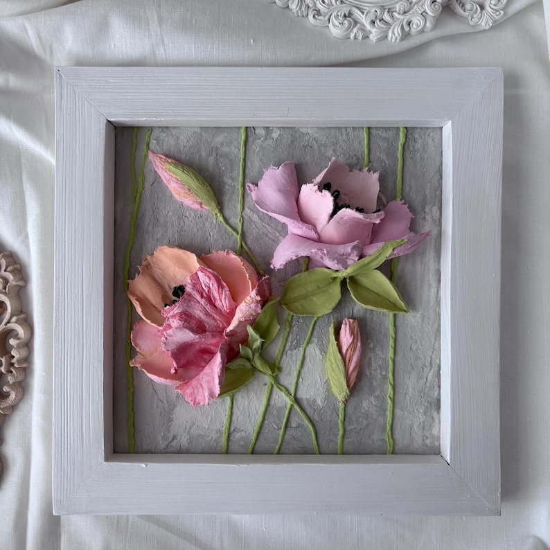 May include: A framed artwork featuring sculpted pink and peach flowers with green leaves and stems against a textured gray background. The flowers have black centers and are set within a white wooden frame, creating a 3D effect.