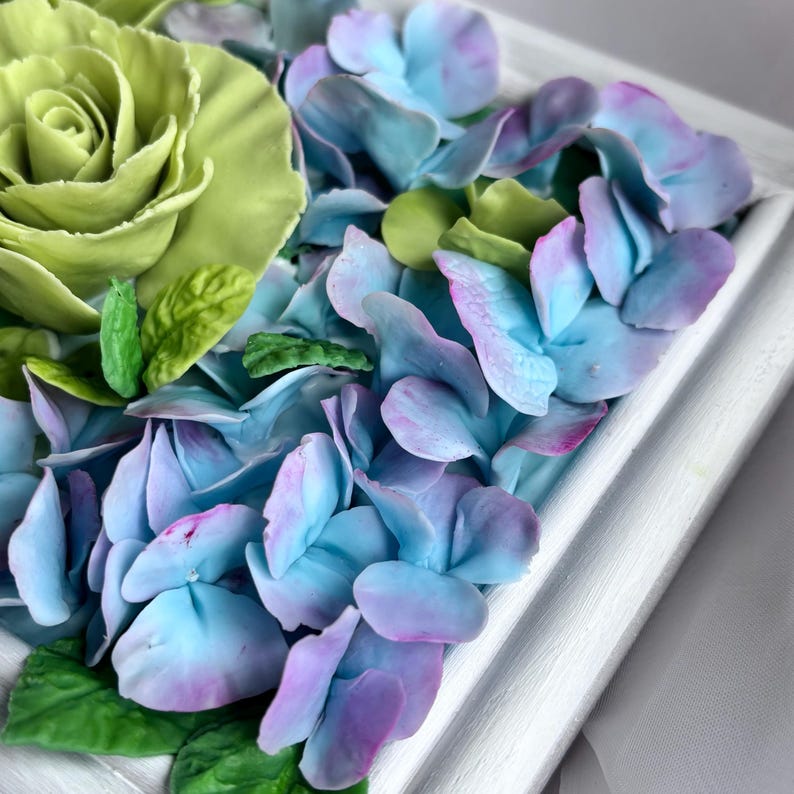 May include: A close-up of a floral arrangement featuring a large, lime green rose and blue and purple hydrangea petals. The flowers are set in a white, square frame, creating a textured, three-dimensional effect. Green leaves are interspersed.