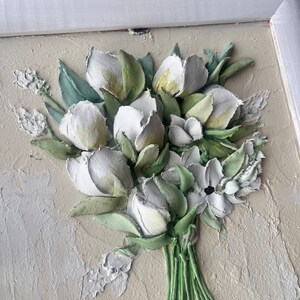 May include: A framed 3D wall art piece featuring a bouquet of white flowers with green leaves. The flowers are sculpted and painted to look realistic.