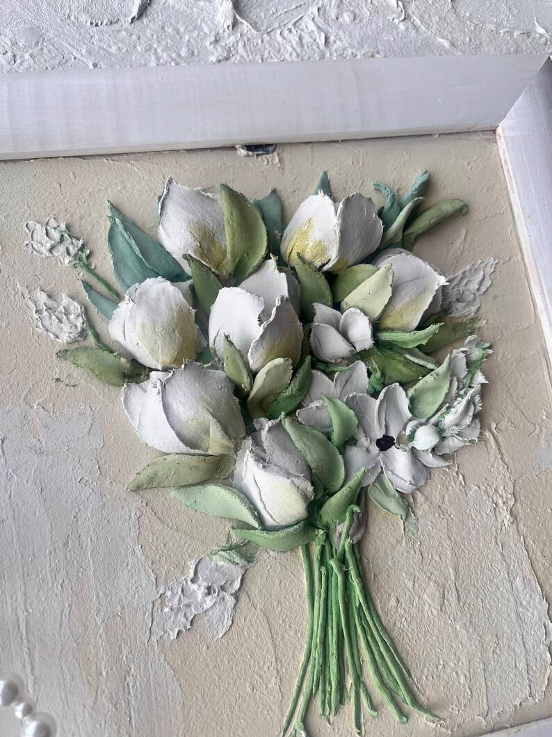 May include: A framed 3D wall art piece featuring a bouquet of white flowers with green leaves. The flowers are sculpted and painted to look realistic.